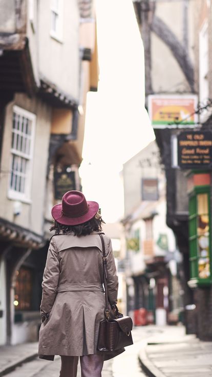 Woman wearing trench coat and pink hat walking through narrow historic street of York, North Yorkshire, England.