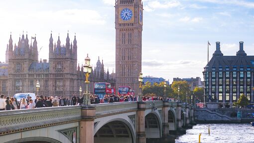 People walk over a busy bridge over a river towards a clock tower and buildings
