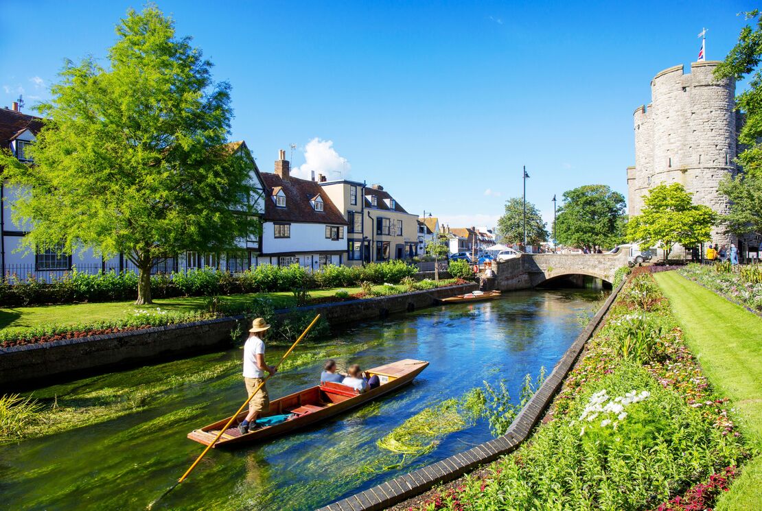 A man and a couple in a punt on a small river surrounded by gardens and houses