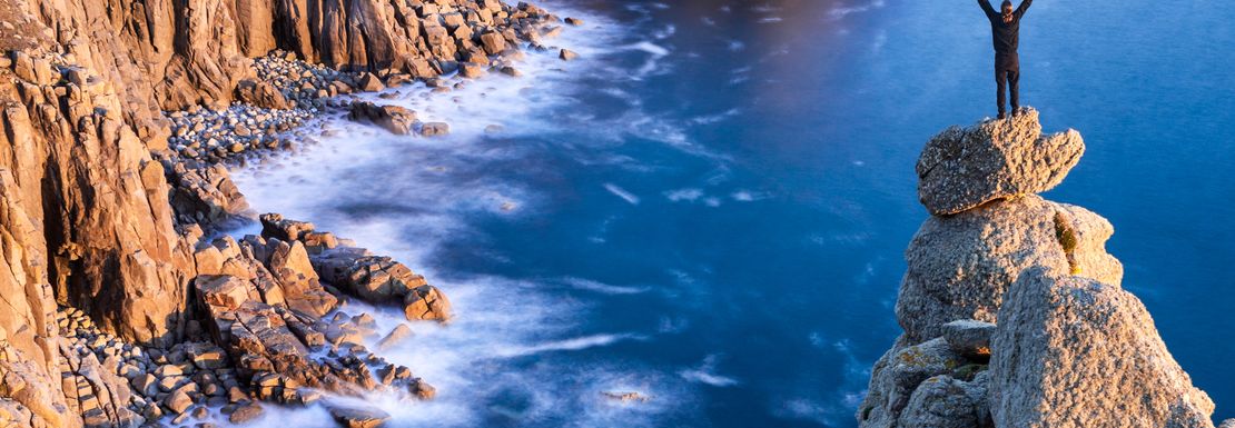 A man stands on a rocky peak looking over the rocky Cornish headland in lands End, Cornwall