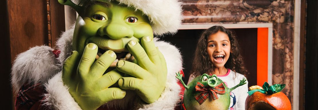 Shrek dressed as Santa covers his mouth as he also smiles and laughs because of a jolly Christmas feeling